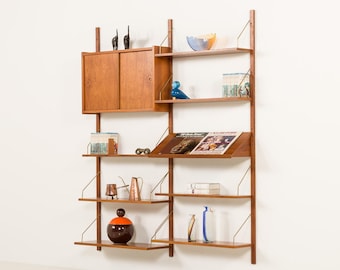 May include: A modular wall-mounted shelving unit made of light brown wood with vertical supports and brass-colored metal brackets. It features a cabinet with sliding doors, multiple open shelves, and a slanted display shelf holding various decorative items.