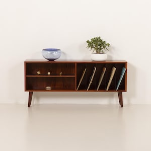 Könnte beinhalten: Ein Mid-Century-Modern-Holzschrank mit abgewinkelten Beinen. Der Schrank hat offene Regale, einen Bereich für Schallplatten und eine kleine Schublade. Eine blaue Glasschale, kleine goldene Kugeln und eine Topfpflanze befinden sich oben.