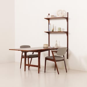 Peut inclure: Un ensemble de salle à manger moderne du milieu du siècle avec une table en bois foncé, deux chaises et une étagère murale. La table rectangulaire est associée à des chaises avec des sièges rembourrés gris. Les étagères exposent des objets décoratifs.