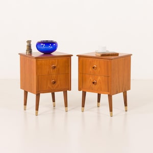 May include: A pair of wooden bedside tables, each with two drawers and tapered legs with brass tips. One table has a blue glass bowl and a brass object. The other has a book and a cup. Each table is approximately 60cm high.