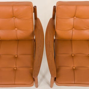 May include: Two brown leather chairs with wooden frames and button tufting. The chairs are positioned side-by-side with the backs facing the camera.