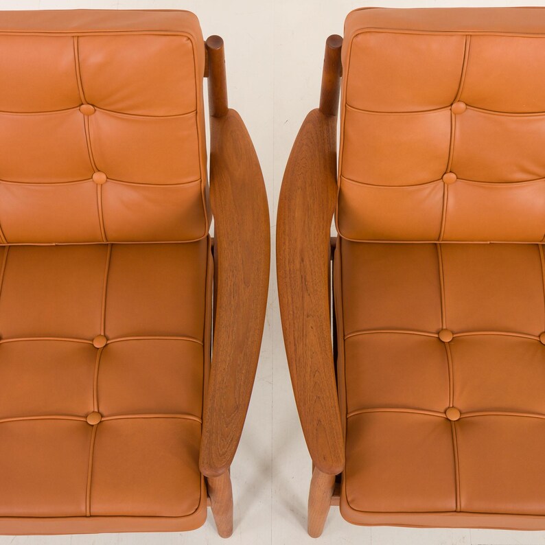 May include: Two brown leather chairs with wooden frames and button tufting. The chairs are positioned side-by-side with the backs facing the camera.