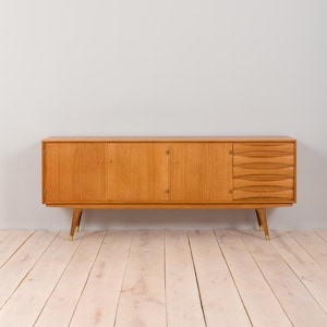 May include: A mid-century modern wooden sideboard with a light brown finish. The cabinet has three doors, four drawers, and angled legs with brass-colored tips. The sideboard is set against a light gray wall and light wood floor.