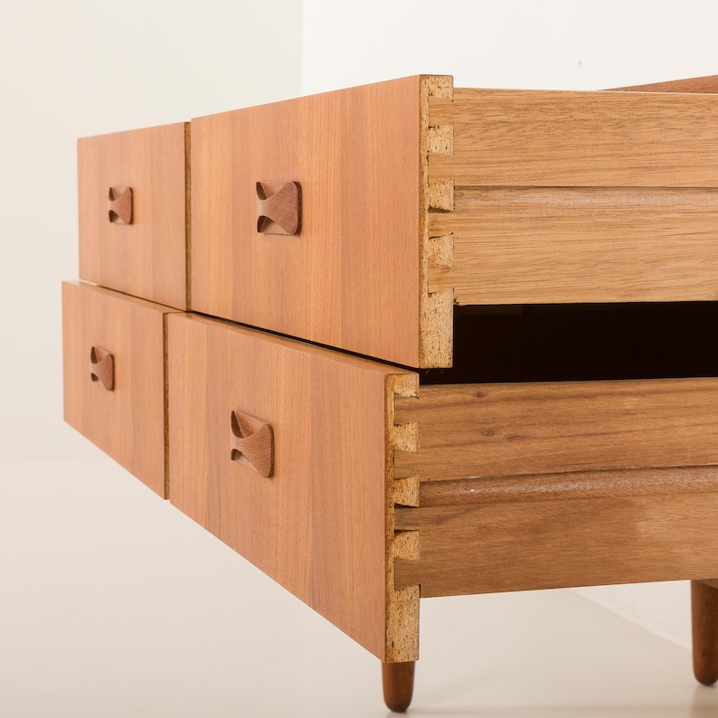 May include: A light brown wooden dresser with four drawers. Two drawers are closed, and two are open, revealing the dovetail joinery. The drawers have small, decorative wooden handles. The dresser is on short, rounded legs.