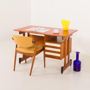 May include: A mid-century modern desk with a red top, a wooden chair with a yellow seat, and a blue glass bottle. The desk has drawers with white and beige stripes. A yellow vase and a cup and saucer are on the desk.