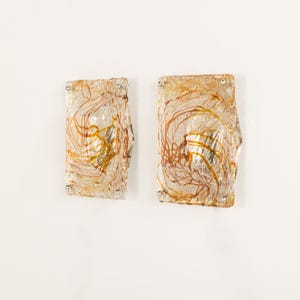 May include: A pair of rectangular wall sconces featuring clear, textured glass panels with swirling amber and brown glass inclusions. Each sconce is secured with four small, metallic silver-toned circular fasteners at the corners of the glass.