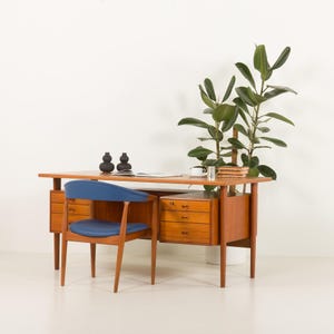 May include: A mid-century modern teak desk with a blue leather chair. The desk has three drawers on the right side and a large open shelf on the left. A plant is in the background.