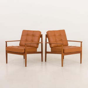 May include: Two brown leather armchairs with wooden frames. The chairs have a tufted design and are positioned side by side on a white background.