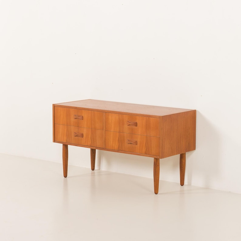 May include: A mid-century modern wooden dresser with four drawers and tapered legs. The dresser is made of light brown wood and features small, decorative wooden handles. The design is simple and elegant, suitable for a bedroom or living space.