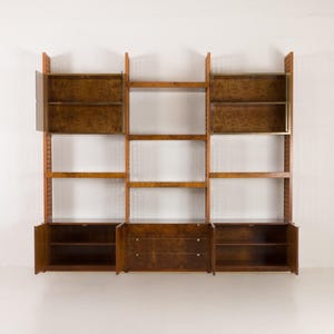 K&ouml;nnte beinhalten: Eine modulare Wand-Einheit aus Holz mit offenen Regalen und Schr&auml;nken. Die Einheit hat eine warme braune Oberfl&auml;che und Messing-Akzente.