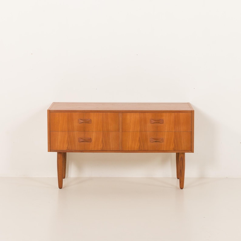 May include: A mid-century modern wooden dresser with four drawers. The dresser is made of light brown wood and has a rectangular shape. The drawers have unique, bow-tie shaped handles. The dresser stands on four tapered legs.