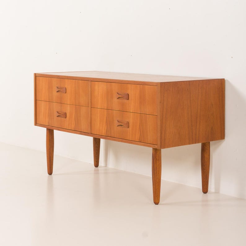 May include: A mid-century modern wooden dresser with four drawers and four tapered legs. The dresser is made of light brown wood and features unique, curved drawer pulls. The design is simple and elegant, perfect for a bedroom or living room.