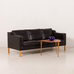 May include: A black leather sofa with three cushions and light wood legs. A small, triangular wooden coffee table with light wood legs sits in front of the sofa. A blue glass bowl is on the table.