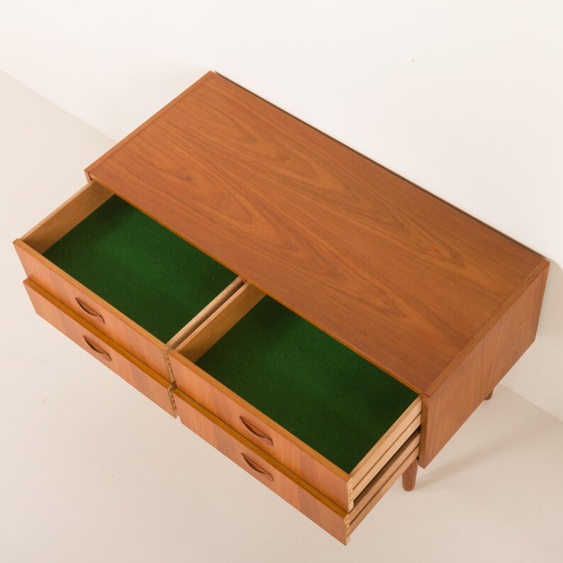 May include: A mid-century modern wooden dresser with four drawers, two of which are open. The drawers have a green felt lining. The dresser is made of light brown wood and has a simple, minimalist design.
