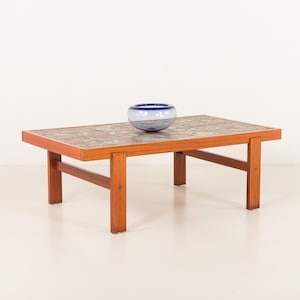 Könnte beinhalten: Ein rechteckiger Couchtisch aus Holz mit einer gefliesten Oberfläche und einer blau-weißen Glasschale. Der Tisch hat eine warme, rötlich-braune Oberfläche und ein schlichtes, stabiles Design. Die geflieste Oberfläche weist ein Muster aus gedämpften Farben auf.