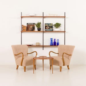 Könnte beinhalten: Ein Wohnzimmer im Retro-Stil mit zwei beigefarbenen Sesseln mit Holzarmlehnen und -beinen, einem kleinen Holztisch und einem Wandregal. Die Regale zeigen Bücher, Pflanzen und Dekorationsgegenstände. Der Raum wirkt hell und luftig.