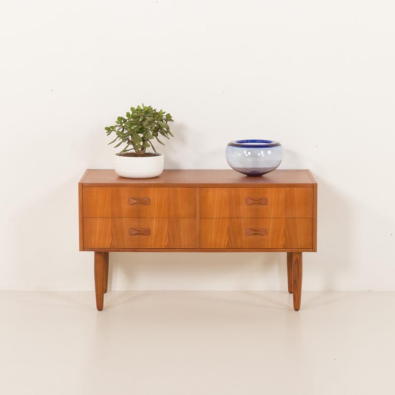 May include: A mid-century modern wooden dresser with four drawers and a light brown finish. The dresser has a rectangular shape and is supported by four tapered legs. A small potted plant and a blue glass bowl sit on top of the dresser.