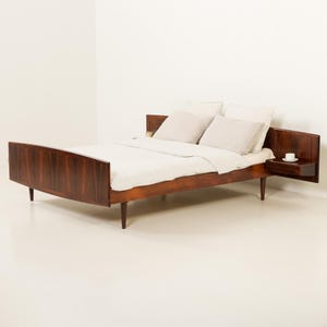 Puede incluir: Una estructura de cama de madera oscura con un patrón de veta prominente, con un cabecero que se extiende hacia mesitas de noche flotantes integradas a ambos lados. La cama está vestida con un edredón blanco y dos almohadas beige claro, sobre patas cónicas.
