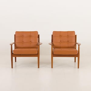 May include: Two brown leather armchairs with button tufting and wooden frames. The chairs are positioned facing each other on a white floor.