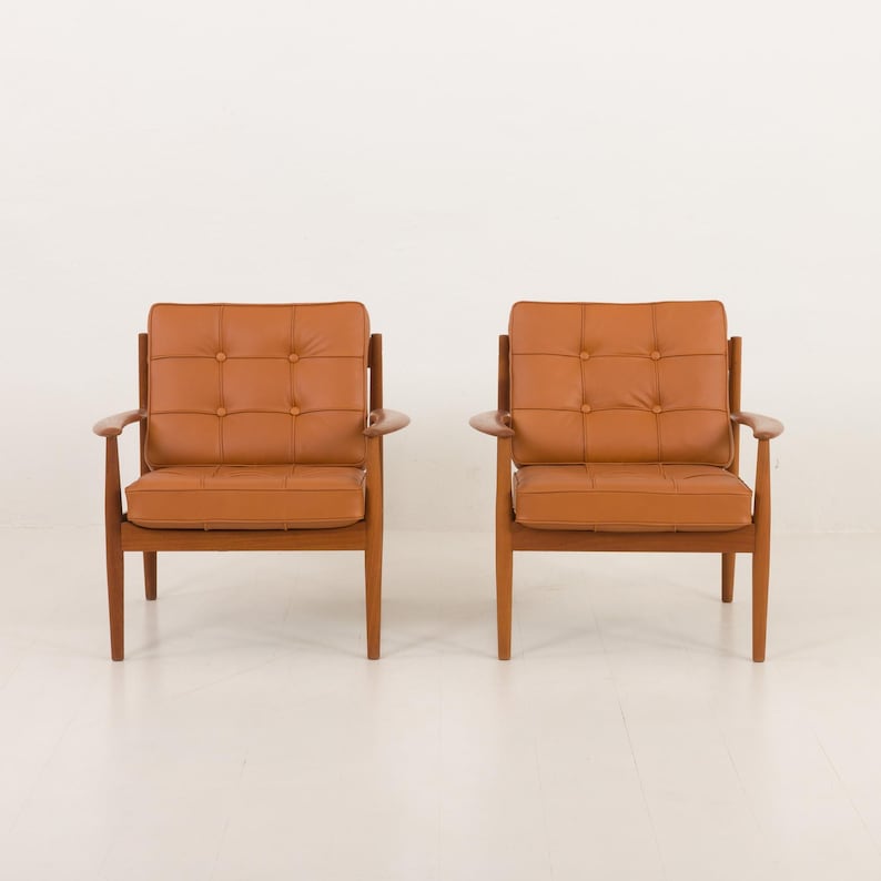May include: Two brown leather armchairs with button tufting and wooden frames. The chairs are positioned facing each other on a white floor.