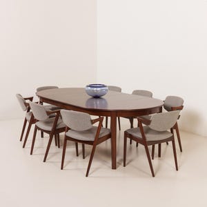 Op de afbeelding: Een donkerbruine houten ovale eettafel met een tussenblad in het midden, vergezeld door acht stoelen met donkerbruine houten frames en lichtgrijze gestoffeerde zittingen en rugleuningen. Een blauwe glazen schaal staat op het tafeloppervlak.