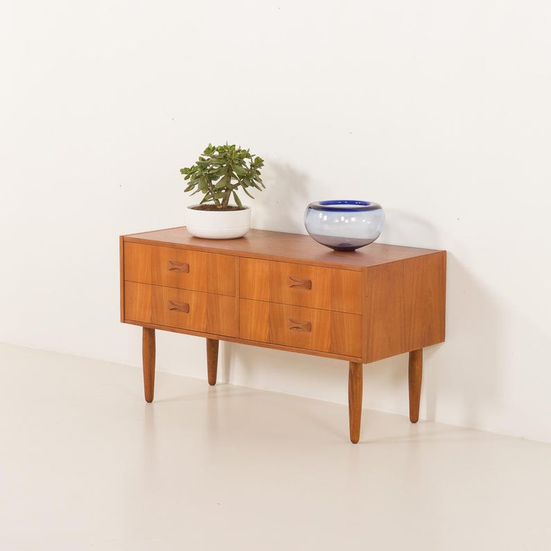 May include: A mid-century modern wooden dresser with four drawers and tapered legs. A small potted plant and a blue and white glass bowl sit on top. The dresser is made of light brown wood.