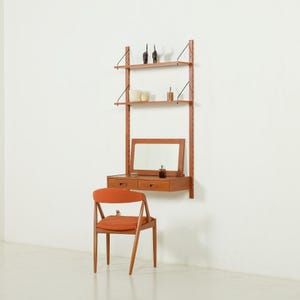 May include: A mid-century modern wall-mounted vanity with a mirror, two drawers, and shelving. The vanity is made of wood and has a warm, reddish-brown finish. An orange chair sits in front of the vanity. Decorative items are on the shelves.