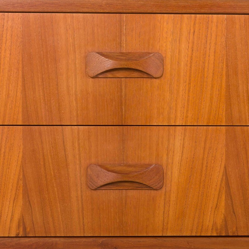 May include: Close-up of a wooden dresser with two drawers. The drawers have a warm, reddish-brown tone with visible wood grain. Each drawer features a unique, curved wooden handle, adding a touch of mid-century modern design.