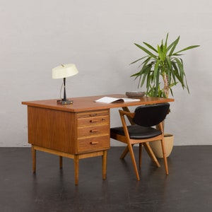 May include: A mid-century modern teak desk with a black leather chair. The desk has four drawers and a lamp with a white shade. The desk is in a room with a white wall and a plant in a brown pot.