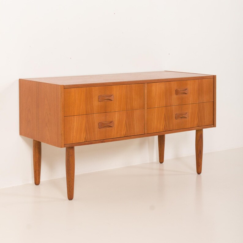 May include: A mid-century modern wooden dresser with four drawers and bow-tie shaped drawer pulls. The dresser is made of light brown wood and has four tapered legs. The dresser is approximately 30 inches wide.