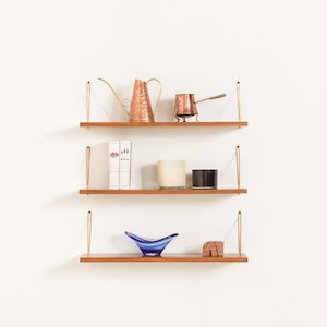 May include: Three-tiered wooden shelves with brass supports, displaying decorative items. The top shelf holds a copper watering can and a small copper pot. The middle shelf features two books, a candle, and a black container. The bottom shelf has a blue glass bowl and a small stone sculpture.
