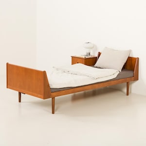 Puede incluir: Una estructura de cama individual de madera de tono medio con cabecero y piecero. La cama está vestida con una sábana bajera gris oscuro, un edredón blanco y una almohada beige claro. Una pequeña mesita de noche de madera con una lámpara blanca está al lado.