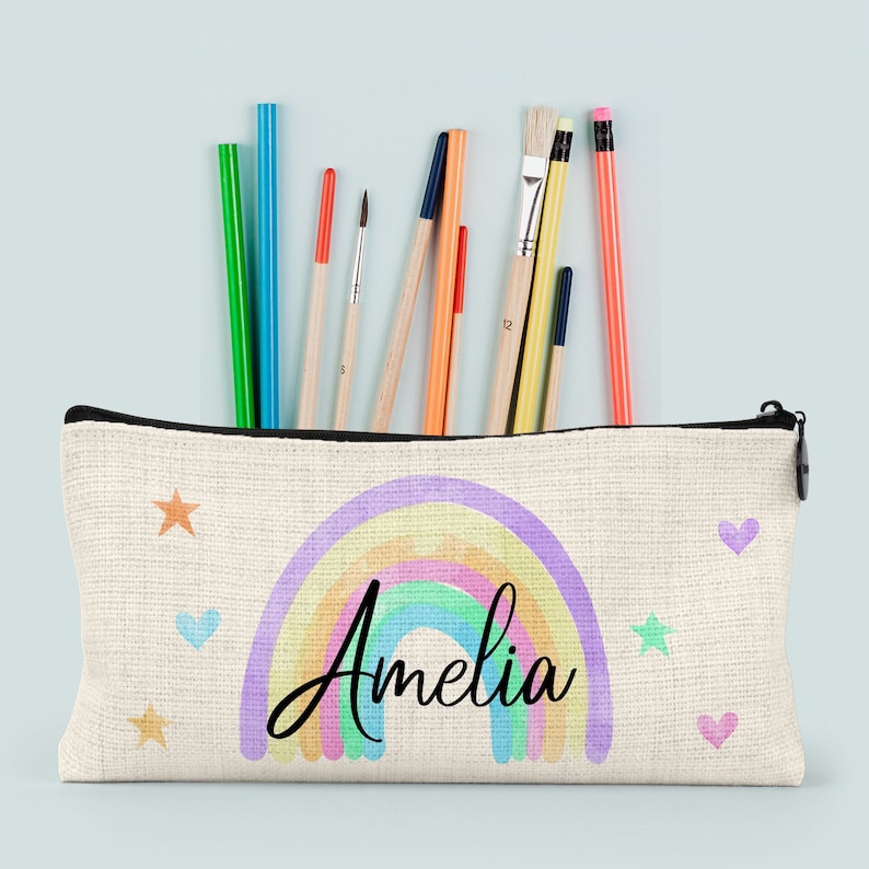 Pu&ograve; includere: Un astuccio per matite in tela bianca con un design arcobaleno e il nome "Amelia" stampato in nero. L'astuccio ha una cerniera e contiene vari materiali artistici, tra cui matite, matite colorate e pennelli.