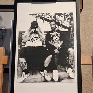 Może przedstawiać: Czarno-biała oprawiona fotografia dwóch osób siedzących na ławce. Zdjęcie jest wyświetlane na drewnianym sztaludze. W tle widoczne są inne oprawione obrazy, a także słowo "MEEDEN" na drewnianym bloku.