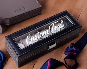 May include: Black leather watch box with a clear glass top. The box has six slots for watches and the text "Custom Text" is printed on the top.