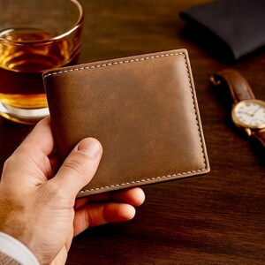Hand holding a brown leather bifold wallet with clean stitching, placed on a dark wooden surface with a glass of whiskey and a brown-band wristwatch in the background for a warm, classy, masculine presentation.