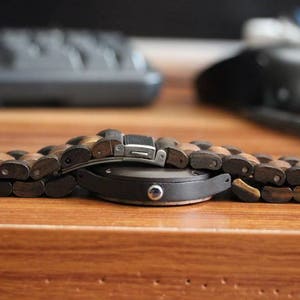 May include: A wristwatch featuring a round black case and a link bracelet constructed from alternating dark and light brown wooden segments. The watch includes a silver-toned metal clasp mechanism visible on the top of the bracelet links.