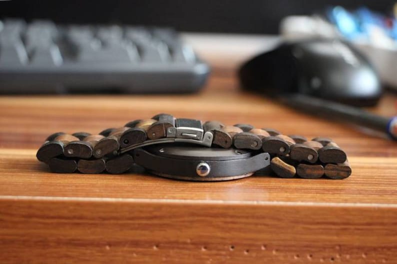 May include: A wristwatch featuring a round black case and a link bracelet constructed from alternating dark and light brown wooden segments. The watch includes a silver-toned metal clasp mechanism visible on the top of the bracelet links.