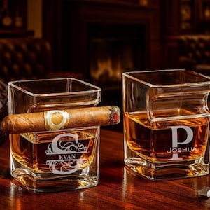 Op de afbeelding: Twee vierkante whiskyglazen met gepersonaliseerde gravures. Eén glas heeft de letter "G" en de naam "EVAN", de andere heeft de letter "P" en de naam "JOSHUA". Een aangestoken sigaar rust op één glas. De glazen zijn gevuld met amberkleurige vloeistof.
