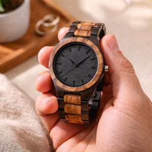 May include: A wristwatch featuring a round face with a solid black dial and thin black hands. The bezel and the link bracelet are constructed from alternating dark brown and light brown wood segments, creating a striped pattern.