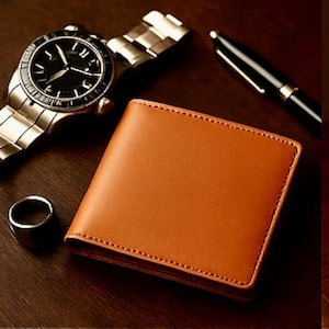 Tan leather bifold wallet displayed on a dark wooden surface beside a silver wristwatch, a black and gold pen, and a metal ring, creating a clean, classy, masculine aesthetic.