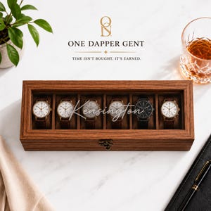 May include: A rectangular wooden watch display case with a glass lid, featuring six individual compartments. Inside, five analog wristwatches with light faces and brown leather straps are arranged alongside one black wristwatch with a black strap.