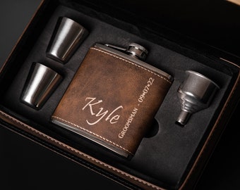 Può includere: Un set regalo con una fiaschetta in pelle marrone incisa con il nome "Kyle" e la data "09-07-22", presentata in una scatola nera. Include anche due bicchierini in acciaio inossidabile e un imbuto.