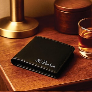Black leather bifold wallet with white cursive engraving reading “K. Paulson,” placed on a warm wooden nightstand beside a brass lamp, a wooden container, and a glass of whiskey for an elegant, masculine presentation.