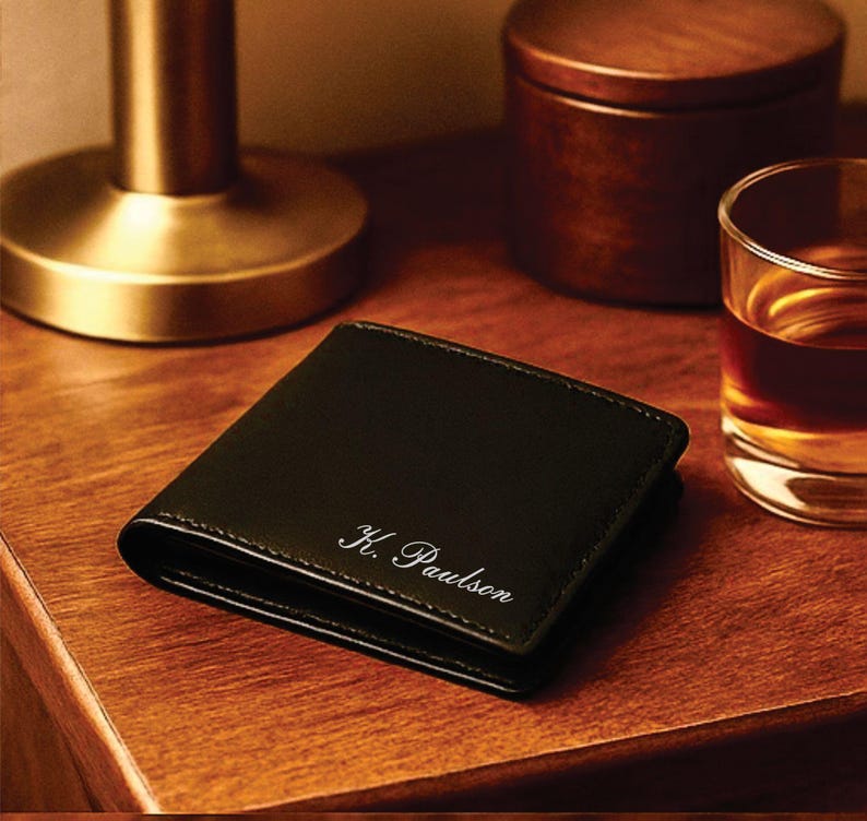 Black leather bifold wallet with white cursive engraving reading “K. Paulson,” placed on a warm wooden nightstand beside a brass lamp, a wooden container, and a glass of whiskey for an elegant, masculine presentation.