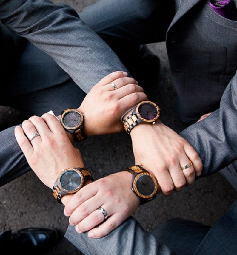 May include: Four wristwatches with dark wooden bands and circular faces featuring dark dials. Each watch is worn on a wrist, and each wrist displays a metallic ring. The watches are arranged in a circular formation, showcasing the wood grain texture and the metallic watch clasps.