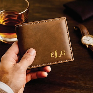Hand holding a brown leather bifold wallet with gold monogram letters “ELG” in the bottom right corner, placed on a dark wooden surface with a glass of whiskey and a brown-band wristwatch in the background for a classy, masculine aesthetic.