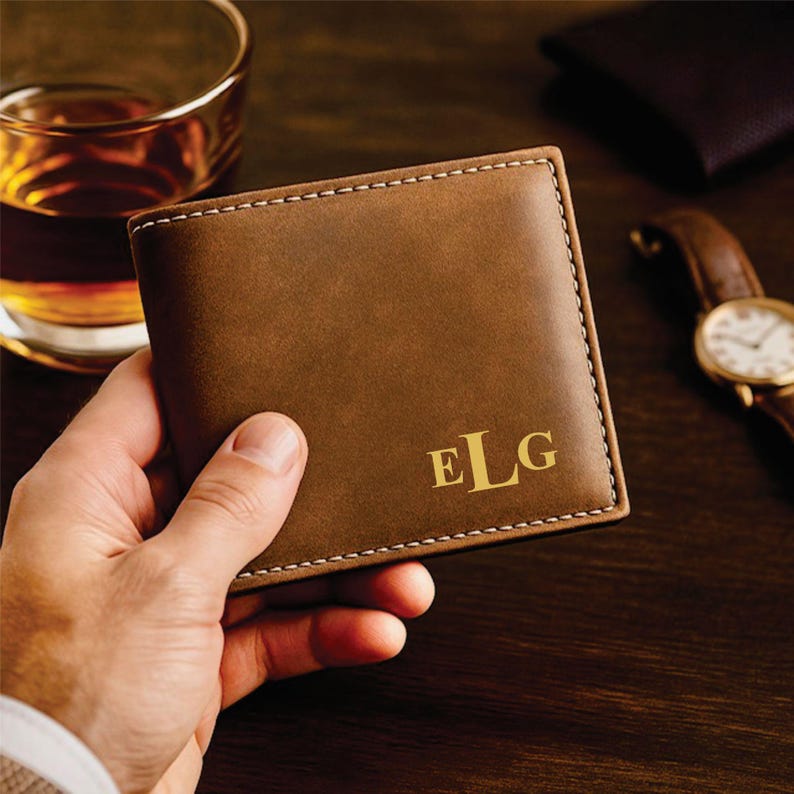 Hand holding a brown leather bifold wallet with gold monogram letters “ELG” in the bottom right corner, placed on a dark wooden surface with a glass of whiskey and a brown-band wristwatch in the background for a classy, masculine aesthetic.
