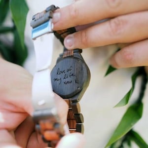May include: A wooden wristwatch with a black face and a brown and black wood band. The back of the watch has a handwritten message that says "love of my life xoxo K".