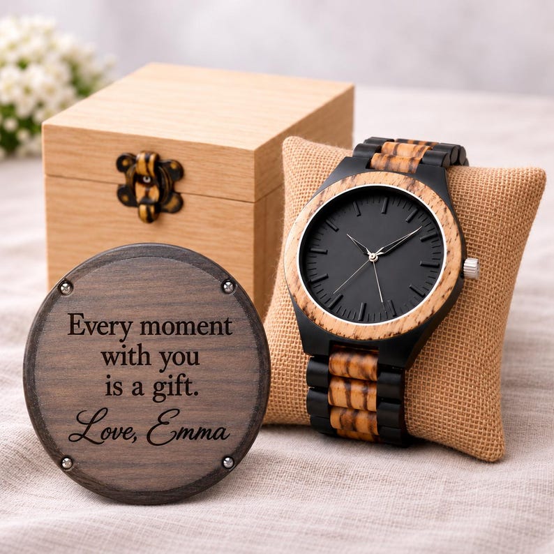 May include: A wooden wristwatch with a black face and a striped wood and black band, displayed on a tan burlap pillow. Beside it is a round wooden plaque engraved with the text: Every moment with you is a gift. Love, Emma. A light wood box is in the back.